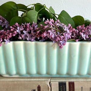 May include: A rectangular, light-blue ceramic planter with vertical ribbed detailing, filled with vibrant purple lilac blooms and green leaves. The planter rests on top of two vintage books.