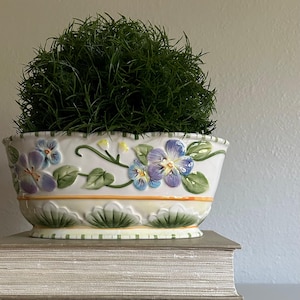 May include: An oval ceramic planter featuring a white base with embossed purple and blue pansy flowers, green leaves, and a scalloped rim. The lower section has a pale yellow band with green leaf motifs and a thin orange stripe, containing a green plant.