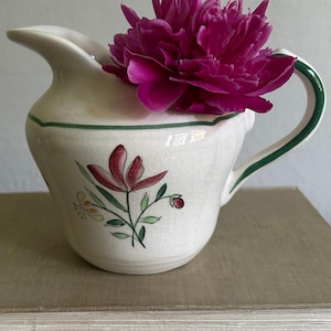 May include: A cream-colored ceramic creamer pitcher with a fine crackle glaze finish, featuring a hand-painted floral motif in red, green, and yellow, a green rim accent, a curved handle, and a single vibrant magenta peony flower inside.