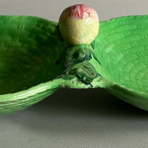 May include: A two-sectioned, green ceramic serving dish with a woven basket texture. A peach-shaped handle sits between the two sections. One side features a painted sprig of cherries. A decorative piece for serving snacks or small items.