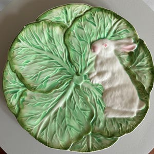 May include: A decorative ceramic plate featuring a white rabbit on a green cabbage leaf design. The plate has a detailed, textured surface with a raised relief of the rabbit and leaf veins. The rabbit has pink ears and red eyes.