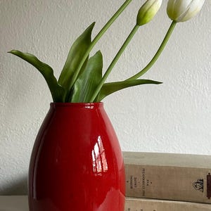 May include: A glossy red vase holds white tulips with green stems and leaves. The vase is set on a white surface, with two vintage books in the background. The books have titles like "The Captive" and "Budding Grove."