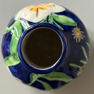 Puede incluir: Un jarr&oacute;n de cer&aacute;mica con fondo azul oscuro, adornado con motivos florales blancos y amarillos y hojas verdes. El jarr&oacute;n tiene forma redonda con interior oscuro y borde azul.