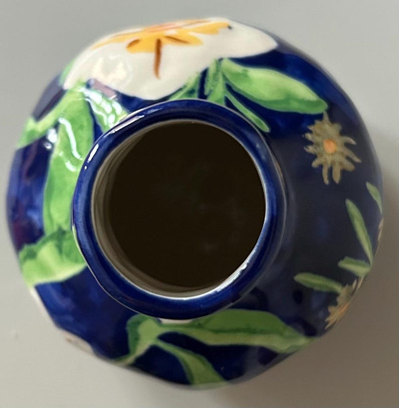 Puede incluir: Un jarr&oacute;n de cer&aacute;mica con fondo azul oscuro, adornado con motivos florales blancos y amarillos y hojas verdes. El jarr&oacute;n tiene forma redonda con interior oscuro y borde azul.