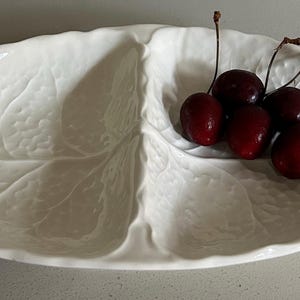 May include: A white ceramic serving dish shaped like a leaf with a divided interior. The dish features a textured surface mimicking natural leaf veins and edges. Five dark red cherries with brown stems are placed in one section of the dish.