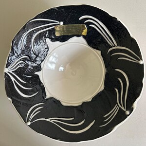 May include: A ceramic serving dish with a central white bowl surrounded by a black rim. The rim features white, stylized leaf designs. A gold label is affixed to the rim. The dish is likely used for serving food or as a decorative piece.