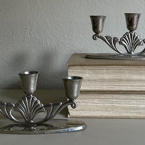 May include: Two silver-toned metal candelabras, each featuring three candle holders arranged on a curved base. The central portion of each candelabra displays a fan-shaped shell motif, with smooth, polished surfaces and a metallic finish.