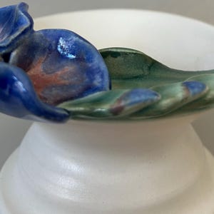 May include: A ceramic pedestal bowl with a white base and a shallow, rounded top. The top is decorated with a blue and green floral design. The blue flower has red accents. The bowl is approximately 6 inches in diameter.