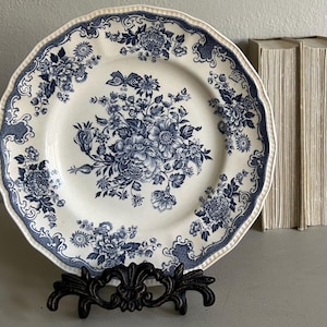 May include: A round ceramic plate with a scalloped edge featuring a blue floral transferware pattern on a cream background. The design includes a central bouquet of flowers and a butterfly, with additional floral motifs along the rim.