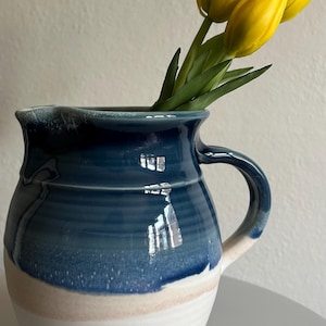 May include: A handmade ceramic pitcher with a blue and white glaze, holding three yellow tulips. The pitcher has a handle and a wide opening. The flowers add a pop of color against the neutral background.