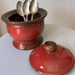 May include: A red ceramic sugar bowl with a lid and three silver spoons. The bowl has a pedestal base and a decorative rim. The lid is red with a brown knob. The spoons are inside the bowl.
