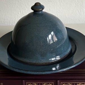 May include: A blue ceramic butter dish with a domed lid and a small knob on top. The dish sits on a matching plate. The glaze has a speckled appearance. The dish is on a dark surface.