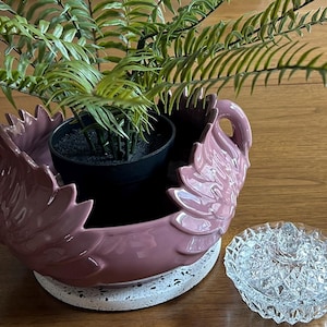 May include: A decorative, mauve-colored ceramic planter with a swan-like design, holding a small potted plant with green foliage. A clear, cut-glass trinket dish sits nearby. The planter rests on a speckled, white base.