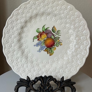 May include: White ceramic plate with a scalloped edge and embossed floral pattern. The center features a colorful fruit and floral design. The plate is displayed on a decorative black metal stand.