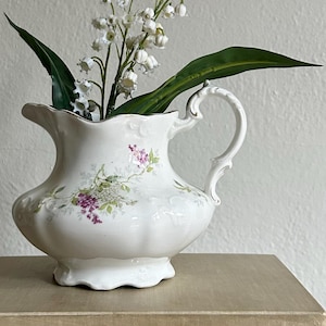 May include: A white porcelain pitcher with a floral design, holding white flowers and green leaves. The pitcher has a curved handle and a scalloped rim. The floral design includes purple and green accents. The pitcher is sitting on top of a book.