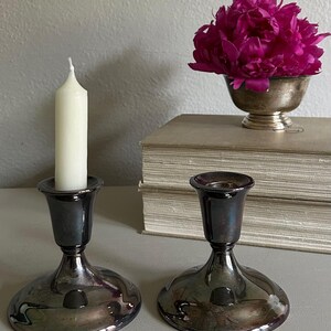 May include: Two polished silver-toned metal candle holders with a wide, circular base and a flared top. One holder contains a single, plain white pillar candle. The metal surface reflects the surrounding environment with a dark, metallic sheen.
