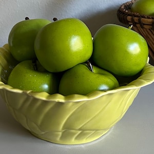 May include: A collection of bright green apples fills a pale yellow, leaf-shaped bowl. The apples have a glossy sheen and dark stems. A woven basket with more green apples is in the background, suggesting a harvest or decorative theme.