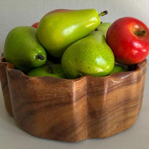 May include: A scalloped wooden bowl filled with artificial fruit, including several bright green pears and a single glossy red apple. The bowl features a natural wood grain finish with a smooth, curved edge design and a deep, rounded interior.