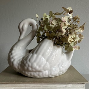 May include: A white ceramic swan-shaped planter featuring a glossy finish and molded feather details. The swan holds a cluster of dried, light green and beige hydrangea flowers within its back, creating a decorative arrangement of natural textures.