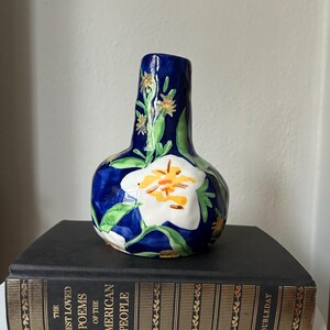 Puede incluir: Un jarr&oacute;n de cer&aacute;mica con una base azul oscuro y un dise&ntilde;o de flor blanca con detalles en amarillo y naranja. El jarr&oacute;n tiene hojas verdes y detalles de flores amarillas. El jarr&oacute;n est&aacute; sobre un libro negro.