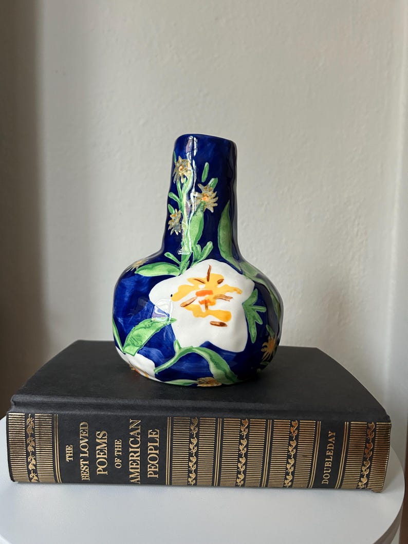 Puede incluir: Un jarr&oacute;n de cer&aacute;mica con una base azul oscuro y un dise&ntilde;o de flor blanca con detalles en amarillo y naranja. El jarr&oacute;n tiene hojas verdes y detalles de flores amarillas. El jarr&oacute;n est&aacute; sobre un libro negro.