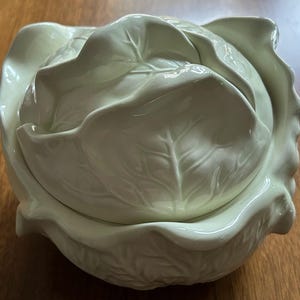 May include: A ceramic cabbage-shaped serving dish with a lid. The dish is a pale green color and has detailed leaf textures. The lid fits snugly, and the overall design is reminiscent of a real cabbage.
