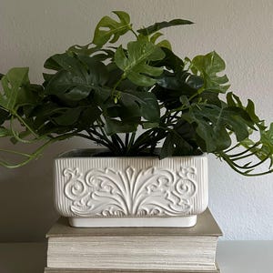 May include: A rectangular white ceramic planter featuring a raised, ornate scrollwork pattern on the front and vertical ribbed detailing on the sides. The planter is filled with a lush, artificial green monstera plant with split leaves.