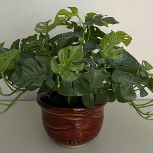 May include: A potted artificial Monstera plant with large, green, split-leaf foliage. The plant is in a brown ceramic pot with a textured surface and decorative details. The plant is a decorative item.