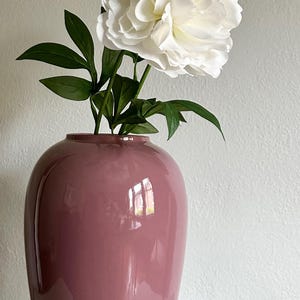 May include: A glossy, mauve-colored vase holds white peony flowers with green leaves. The vase has a rounded shape and a smooth, reflective surface. The flowers are large and full, with multiple layers of petals.