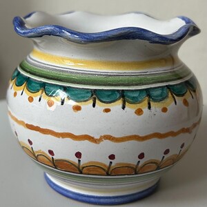 May include: A white ceramic vase with a ruffled rim and a blue trim. The vase is decorated with horizontal bands of yellow, green, and orange, with small floral and dot patterns. The base also has a blue trim.