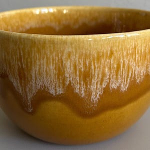 May include: A ceramic bowl with a glossy, glazed finish. The bowl features a gradient of colors, transitioning from a light yellow at the top to a deep amber at the base. White streaks add texture to the design.