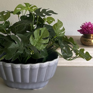 May include: A light gray, oval-shaped ceramic planter with a fluted, scalloped rim design. The planter contains a lush, artificial green monstera plant with split leaves. The piece features a glossy finish with subtle white highlights along the edges.