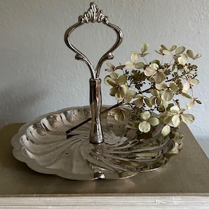 May include: A silver-toned metal serving tray with a scalloped edge and a central vertical handle featuring a decorative loop at the top. A dried hydrangea flower stem with pale green and cream-colored petals rests across the reflective surface.