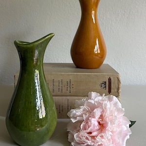 May include: Two ceramic vases, one green and one orange, sit atop a stack of vintage books. A large, light pink peony flower rests in front of the books. The vases have a unique, curved design.