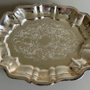 May include: A silver-colored, antique serving tray with a scalloped edge. The tray features an ornate, engraved floral pattern in the center. The tray's surface reflects light, highlighting its detailed design and aged patina.