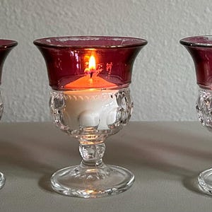 May include: Five vintage glass candle holders. Each has a clear, patterned base and a deep red bowl. One candle holder contains a lit white candle. The candle holders are arranged in a row on a light-colored surface.