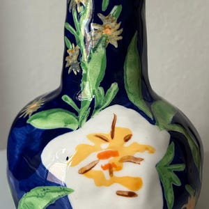 Puede incluir: Un jarr&oacute;n de cer&aacute;mica con un fondo azul oscuro y una gran flor blanca con detalles amarillos y naranjas. Hojas verdes y flores amarillas m&aacute;s peque&ntilde;as est&aacute;n pintadas en el jarr&oacute;n. El jarr&oacute;n tiene un cuerpo redondeado y un cuello estrecho.