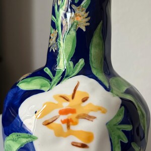 Puede incluir: Un jarr&oacute;n de cer&aacute;mica pintado a mano con un fondo azul oscuro y dise&ntilde;os florales. El jarr&oacute;n presenta una gran flor blanca con detalles amarillos y naranjas, junto con hojas verdes y flores amarillas m&aacute;s peque&ntilde;as. El jarr&oacute;n mide unos 18 cm de alto.