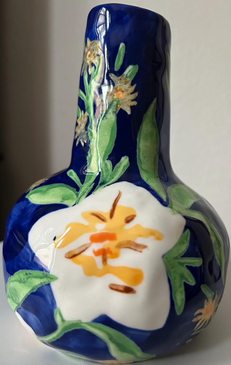 Puede incluir: Un jarr&oacute;n de cer&aacute;mica pintado a mano con un fondo azul oscuro y dise&ntilde;os florales. El jarr&oacute;n presenta una gran flor blanca con detalles amarillos y naranjas, junto con hojas verdes y flores amarillas m&aacute;s peque&ntilde;as. El jarr&oacute;n mide unos 18 cm de alto.