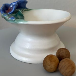 May include: A white ceramic pedestal bowl with a blue and green floral accent. Three small wooden spheres are in front of the bowl. The bowl has a wide, shallow opening and a flared base. The floral accent features blue petals and green leaves.