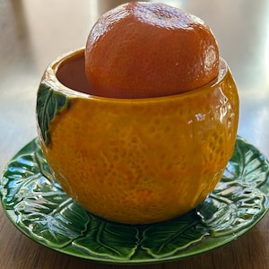 May include: A ceramic orange-shaped bowl with a tangerine inside, resting on a green leaf-shaped saucer. The bowl is textured and has a glossy finish. The saucer is a vibrant green color.