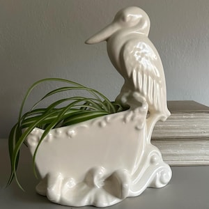 May include: A cream-colored ceramic planter shaped like a boat with a pelican perched on the stern. The vessel features molded wave-like details at the base and holds a green spider plant with long, thin, variegated leaves arching over the sides.