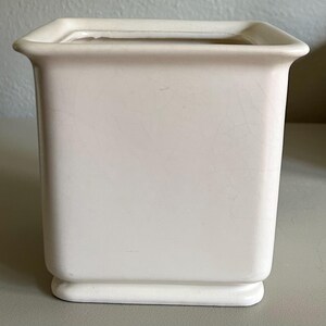 May include: A square, off-white ceramic planter with a slightly flared rim. The planter has a rectangular base and a smooth, matte finish. The interior is a darker shade of beige. Cracks are visible on the surface.