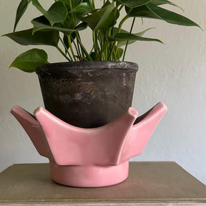 May include: A glossy pink ceramic plant pot holder with a star-shaped rim and a cylindrical base. A dark, weathered, round terracotta pot containing a green leafy plant with heart-shaped foliage sits inside the pink ceramic holder.