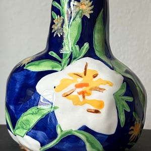 Puede incluir: Un jarr&oacute;n de cer&aacute;mica pintado a mano con un fondo azul oscuro y dise&ntilde;os florales blancos y amarillos. El jarr&oacute;n tiene forma bulbosa con cuello estrecho y est&aacute; decorado con hojas verdes.