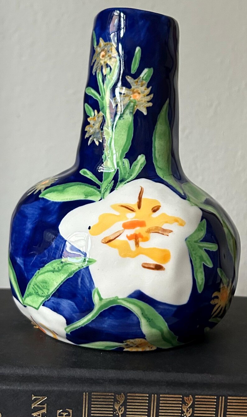 Puede incluir: Un jarr&oacute;n de cer&aacute;mica pintado a mano con un fondo azul oscuro y dise&ntilde;os florales blancos y amarillos. El jarr&oacute;n tiene forma bulbosa con cuello estrecho y est&aacute; decorado con hojas verdes.
