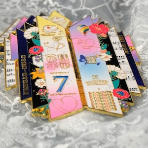 May include: A collection of colorful metal pins with gold trim. The pins feature floral designs, musical notes, and text including "Butter", "LY", "7", and "BE". The color palette includes pink, blue, yellow, and black.