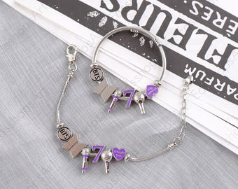 May include: Two silver-toned charm bracelets featuring metallic charms including a circular BTS logo, a geometric shape, a purple and silver microphone, the number seven in purple, and a purple heart charm with the text I Purple You.