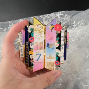 May include: A colorful pin featuring a stylized design with floral accents and the text "BTS2013.6.13-2023.6.13" and "ARMY FOREVER". The pin has a gold border and includes the number "7" and the word "BE".