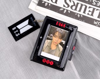May include: A black plastic photo card holder featuring a portrait of a person with dark hair wearing a light-coloured shirt. The frame is decorated with red BTS text, white star accents, and a black tag with the white BTS logo attached by a metal chain.